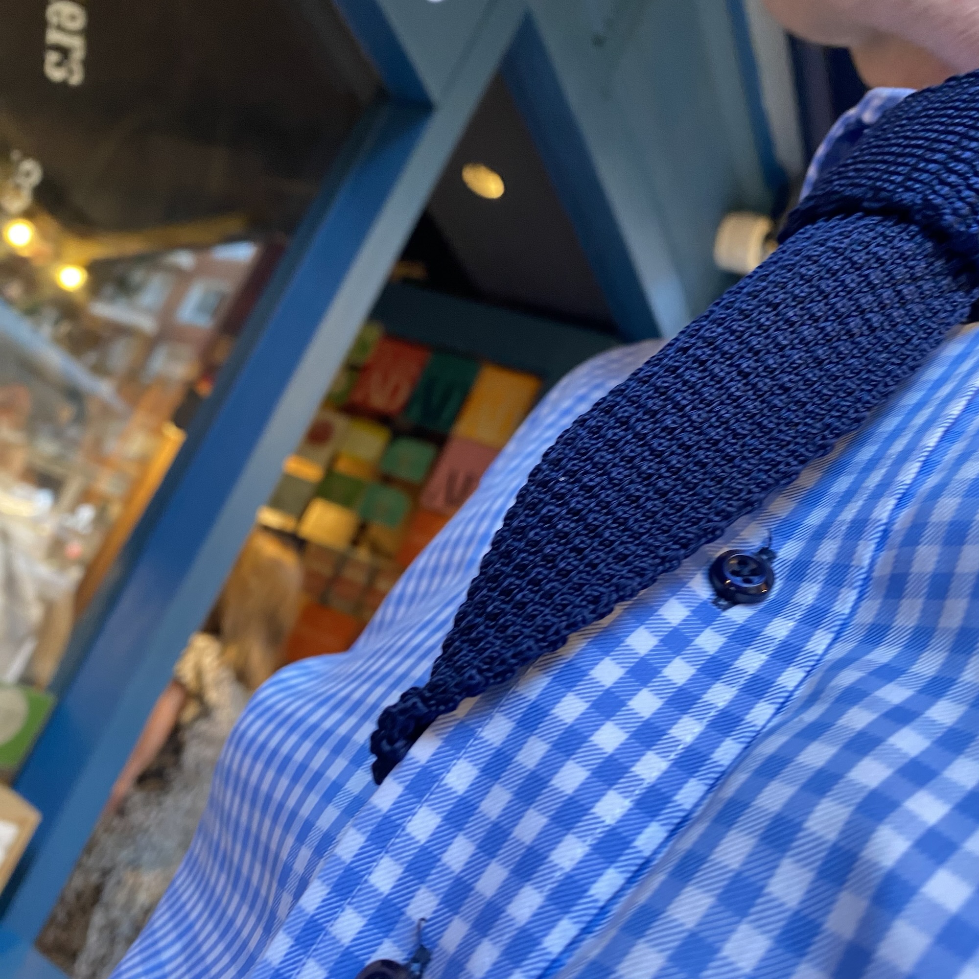 A blue check shirt is worn with a dark knitted tie tucked loosely between the shirt buttons at a casual, angled close-up.