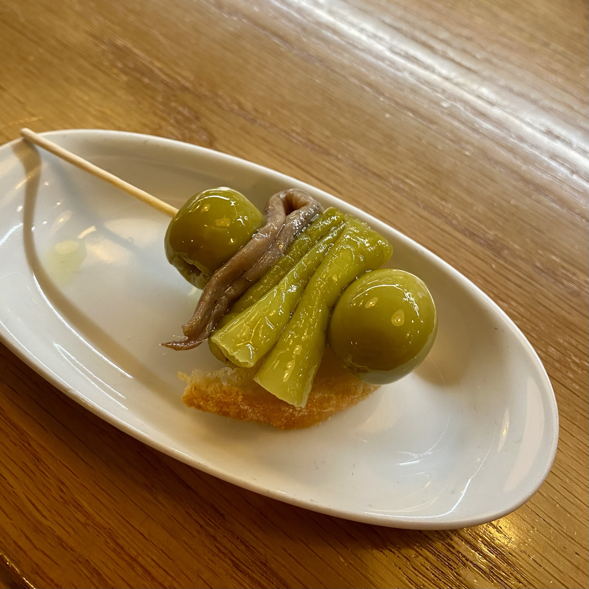 A white oval dish holds green olives, anchovy fillets and sliced pickled peppers on a wooden table.