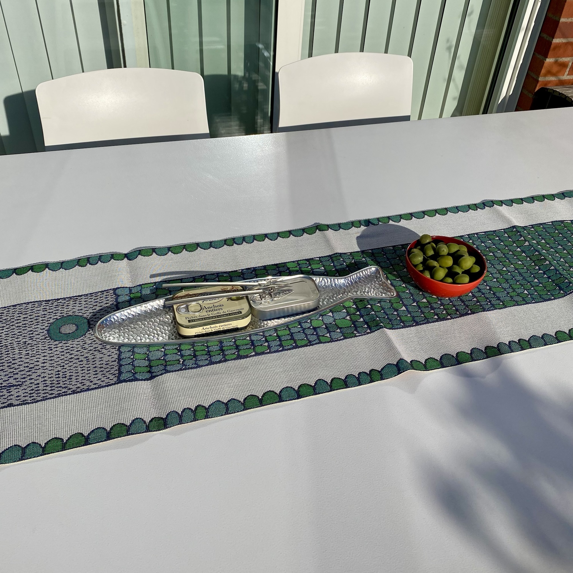 A woven table runner with Stig Lindberg’s fish motif stretches across a sunlit table beside anchovies and olives.