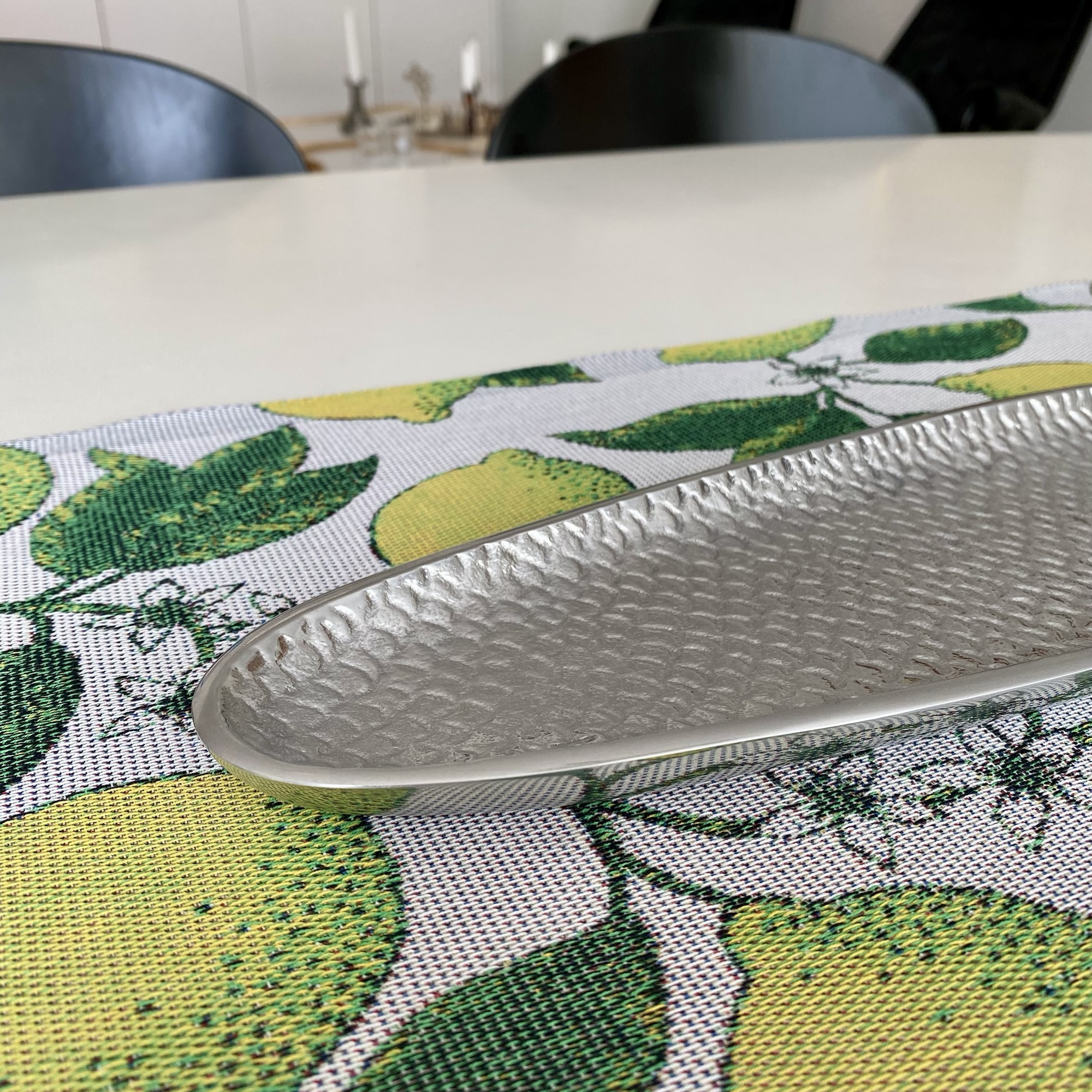 A hammered silver Fish & Fish platter rests on a lemon-patterned runner beside a pale Superellipse table and dark chairs.