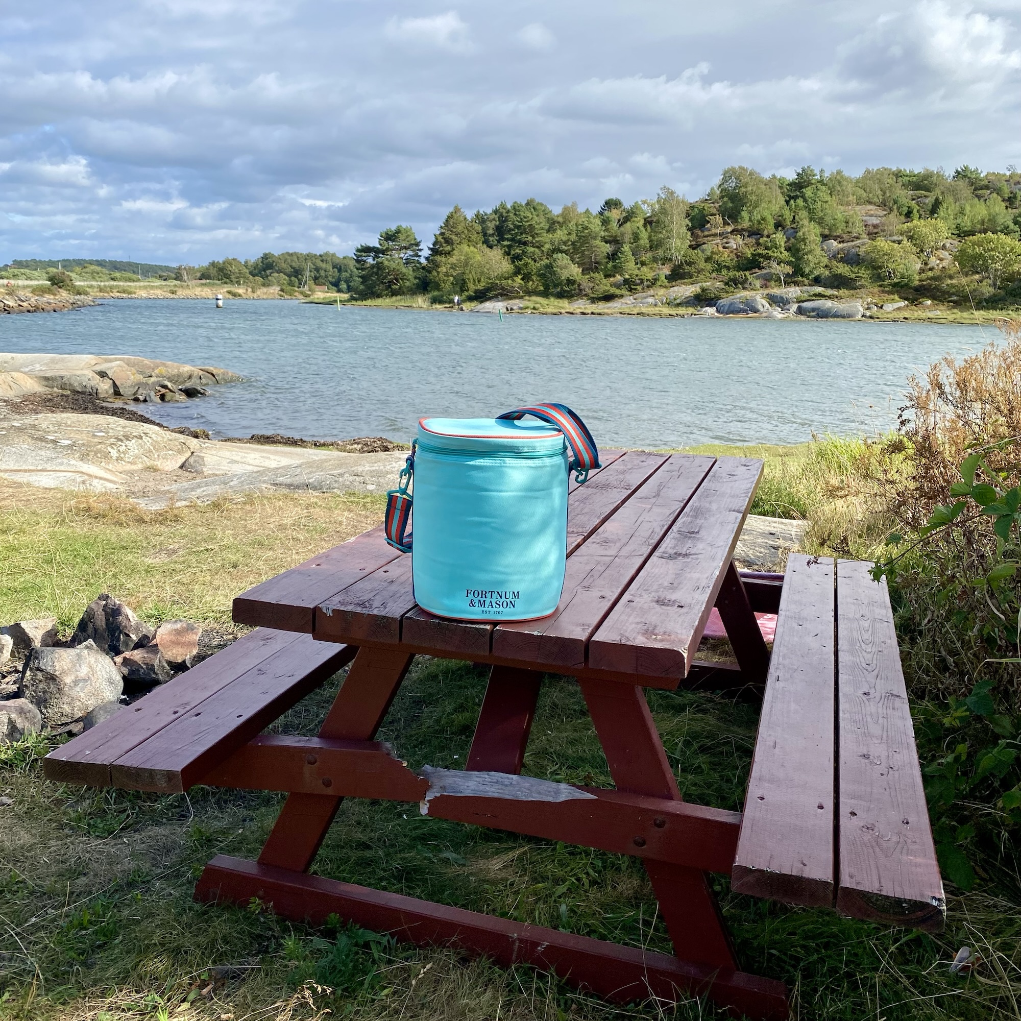A turquoise Fortnum & Mason picnic bag rests on a wooden picnic table beside a calm rocky bay and wooded shoreline.