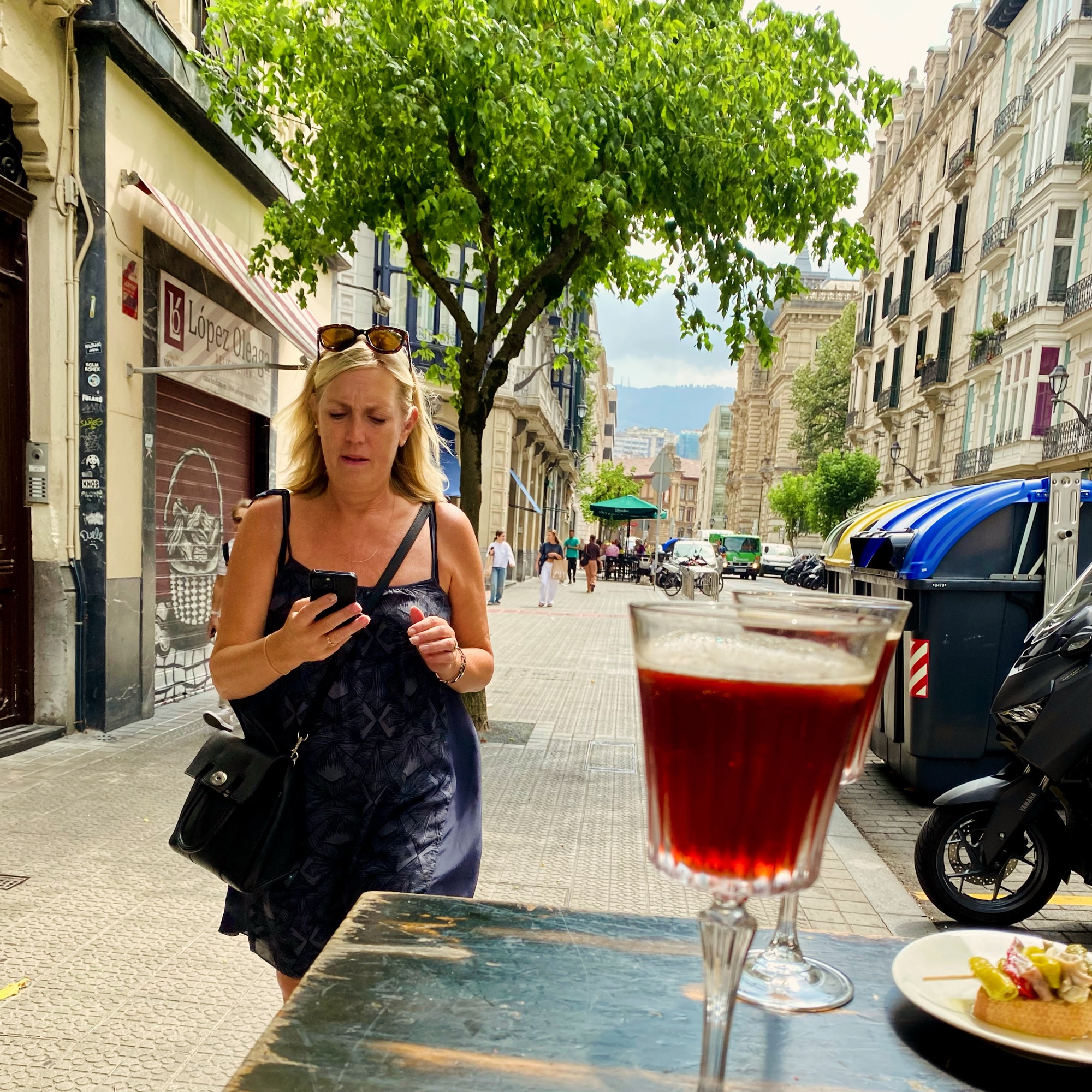 A blonde woman checks her phone on a city street, with a red cocktail glass and tapas in the foreground.