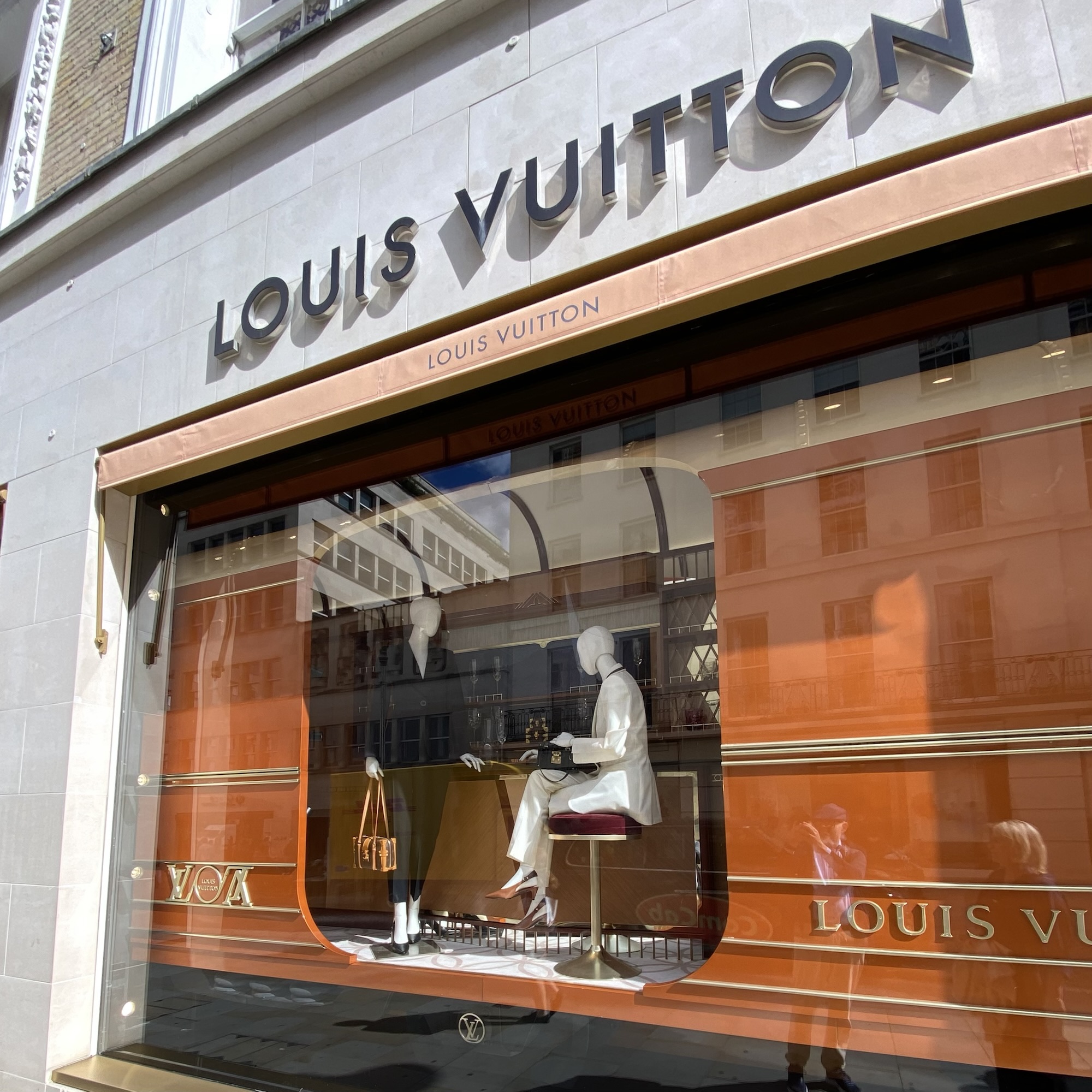 A Louis Vuitton storefront displays mannequins and handbags in an orange Art Deco window with reflective glass.