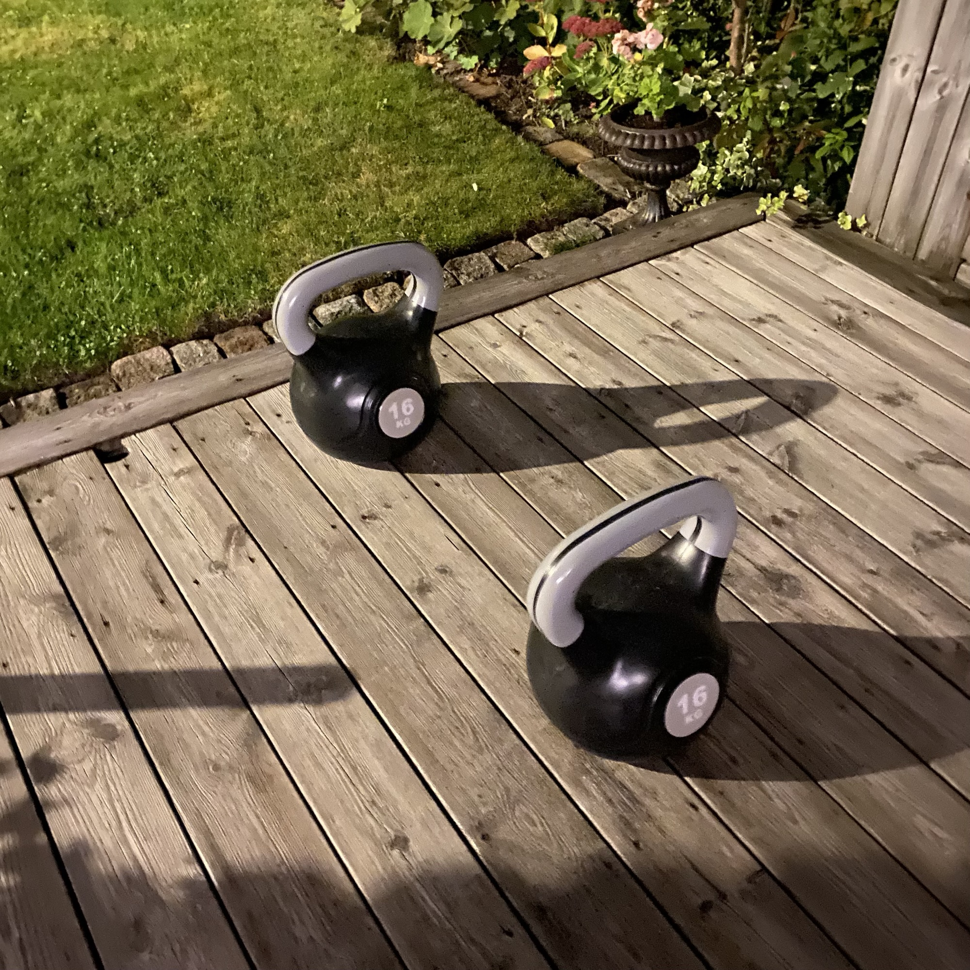 Two 16 kg kettlebells sit on a wooden garden deck beside a lawn and flower bed as autumn evenings grow darker.