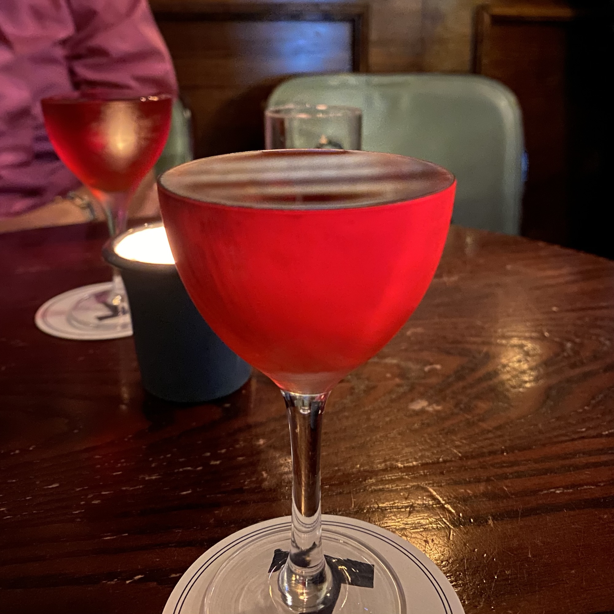 A red Negroni fills a stemmed glass to the rim on a wooden bar at Bar Termini, with another drink and candle behind it.