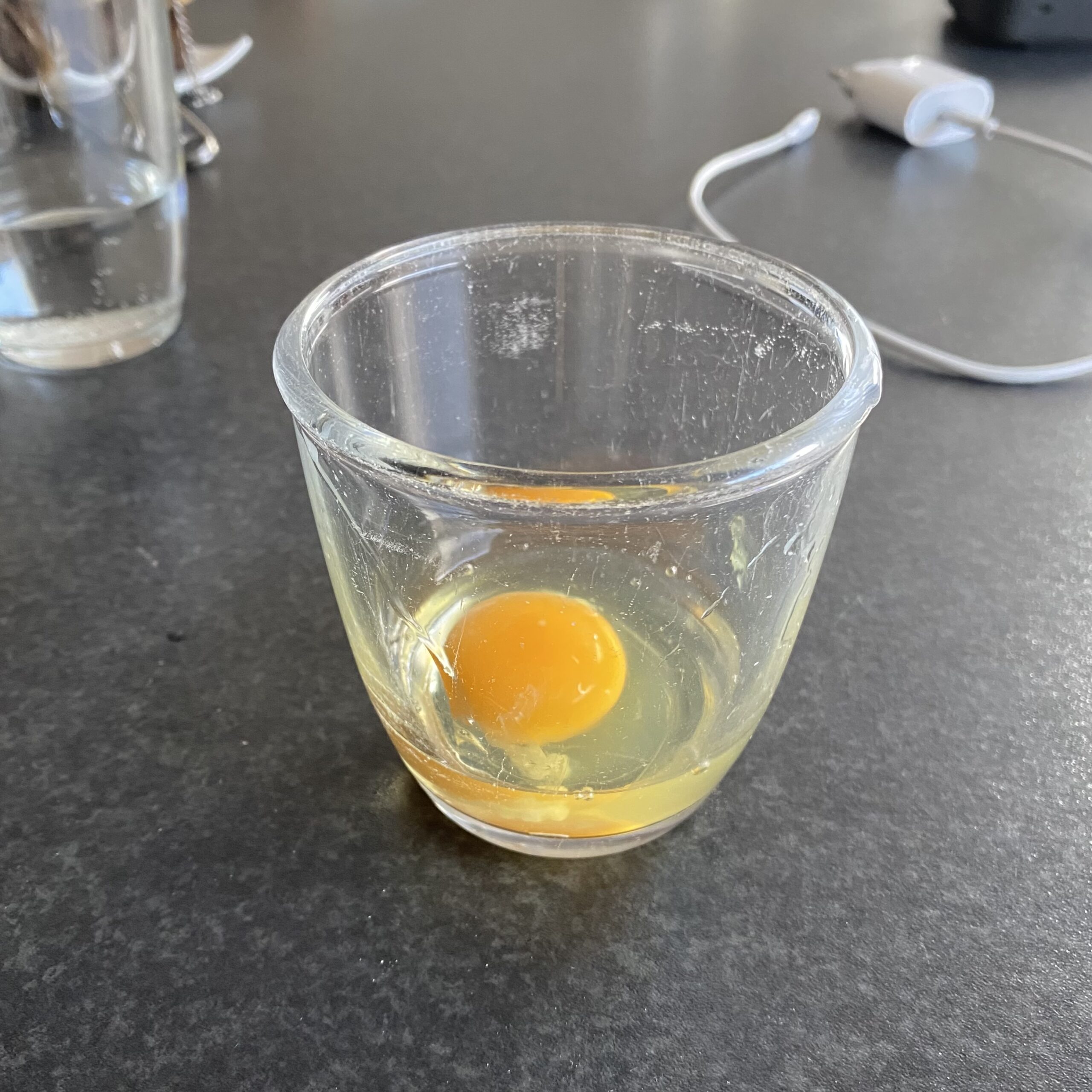 A raw egg with an orange yolk rests in a clear glass cup on a dark counter, part of the business lunch before the gym.
